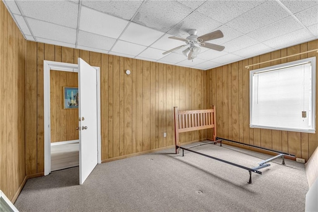 unfurnished bedroom with a ceiling fan, a drop ceiling, carpet floors, wooden walls, and baseboards