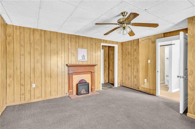 unfurnished living room featuring wooden walls, baseboards, carpet floors, a fireplace, and a ceiling fan