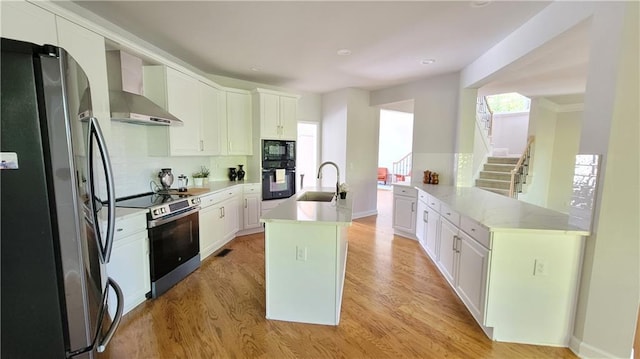 kitchen with wall chimney range hood, sink, appliances with stainless steel finishes, white cabinetry, and a center island with sink