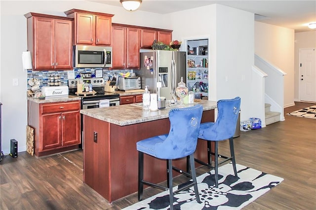 kitchen with a kitchen bar, an island with sink, stainless steel appliances, decorative backsplash, and dark hardwood / wood-style floors