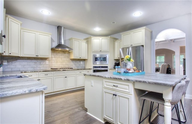 116 Chorley Run, Ellenwood, GA 30294 - kitchen featuring wall chimney range hood, appliances with stainless steel finishes, arched walkways, light stone countertops, and light wood-style flooring