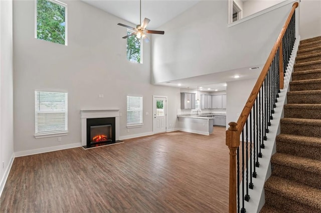 unfurnished living room featuring ceiling fan, a glass covered fireplace, wood finished floors, a towering ceiling, and stairway