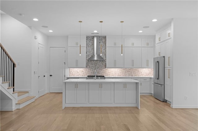 kitchen with high end refrigerator, wall chimney exhaust hood, pendant lighting, and an island with sink