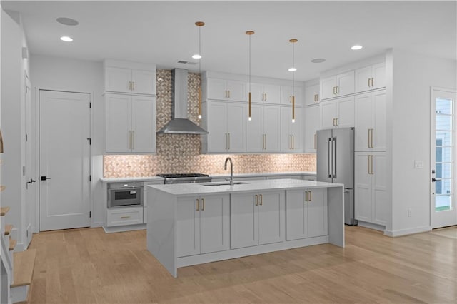 kitchen featuring high end fridge, wall chimney exhaust hood, a kitchen island with sink, sink, and decorative light fixtures