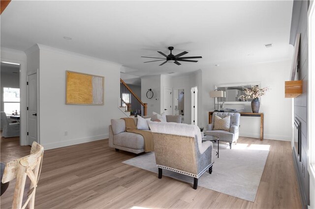 living room featuring a ceiling fan, baseboards, light wood finished floors, stairs, and crown molding