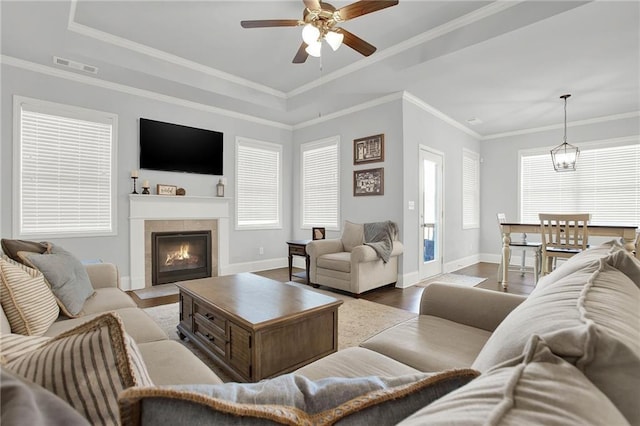 living room featuring a tile fireplace, ceiling fan with notable chandelier, ornamental molding, a tray ceiling, and wood-type flooring