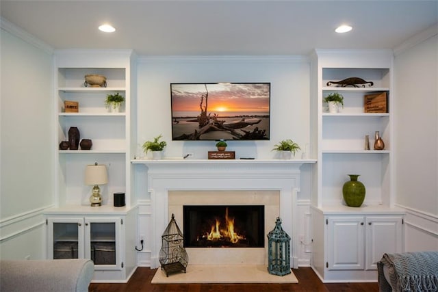 living area with built in features, dark wood-type flooring, a warm lit fireplace, crown molding, and recessed lighting