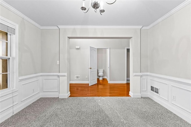 carpeted spare room featuring a wainscoted wall, a decorative wall, visible vents, and crown molding