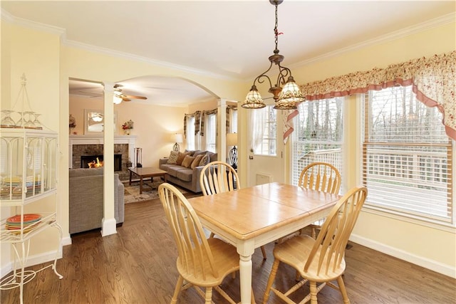 dining space with ornamental molding, ceiling fan with notable chandelier, a warm lit fireplace, wood finished floors, and arched walkways