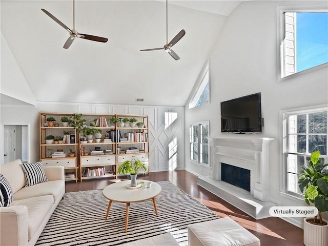 living area featuring a fireplace with raised hearth, visible vents, a ceiling fan, wood finished floors, and high vaulted ceiling