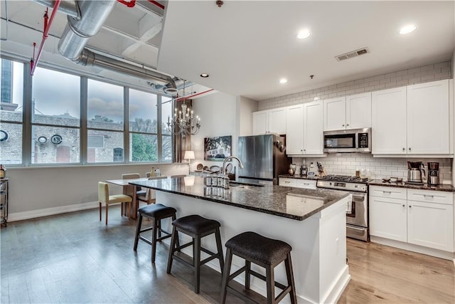 kitchen with a center island with sink, stainless steel appliances, visible vents, white cabinetry, and dark stone counters