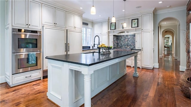 kitchen featuring white cabinetry, stainless steel double oven, ornamental molding, and a center island with sink