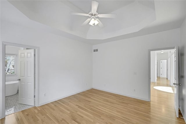 unfurnished bedroom featuring ceiling fan, a raised ceiling, ensuite bath, and light wood-type flooring