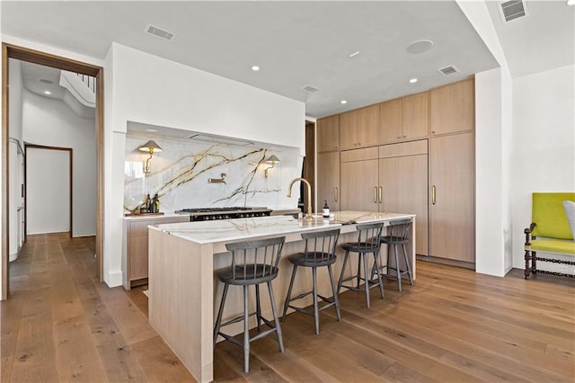 kitchen with an island with sink, a breakfast bar area, decorative backsplash, light stone counters, and light hardwood / wood-style flooring