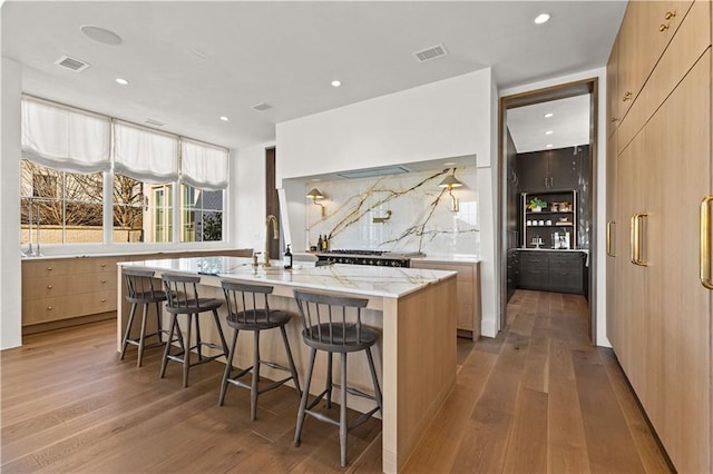 kitchen with hardwood / wood-style floors, an island with sink, a breakfast bar area, backsplash, and light stone counters