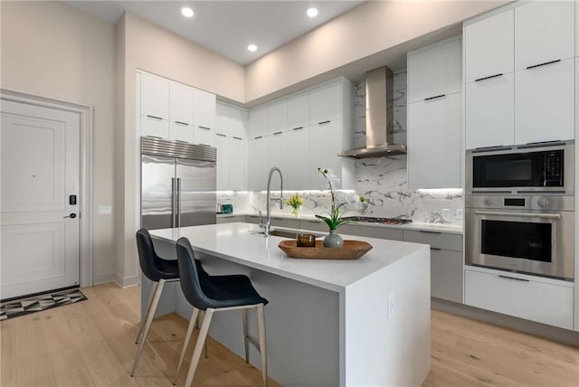 kitchen featuring white cabinets, wall chimney range hood, decorative backsplash, an island with sink, and a breakfast bar area