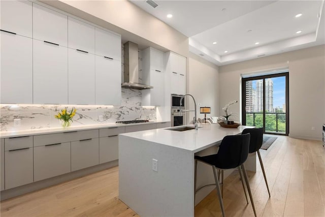 kitchen with light hardwood / wood-style flooring, wall chimney exhaust hood, decorative backsplash, an island with sink, and a kitchen bar