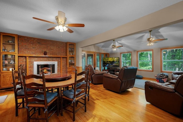 dining area with light wood-type flooring, a textured ceiling, ceiling fan, and a brick fireplace