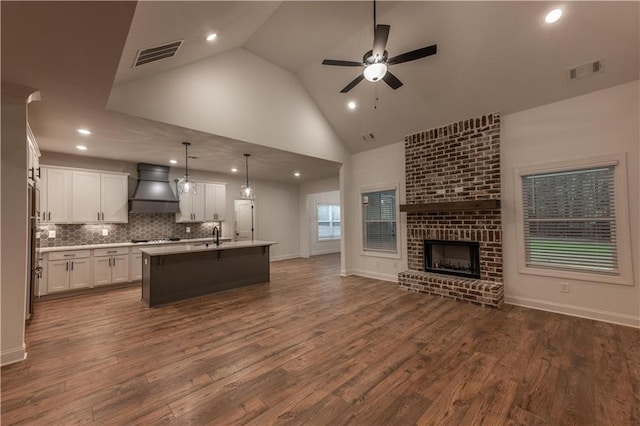 kitchen with premium range hood, a brick fireplace, dark wood-type flooring, pendant lighting, and an island with sink