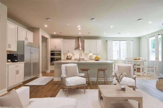 kitchen with white cabinets, wood-type flooring, an island with sink, and wall chimney range hood