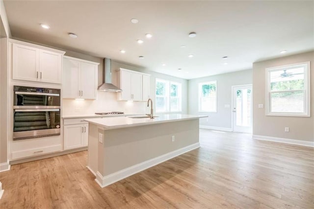 kitchen with double oven, white cabinetry, an island with sink, and wall chimney range hood