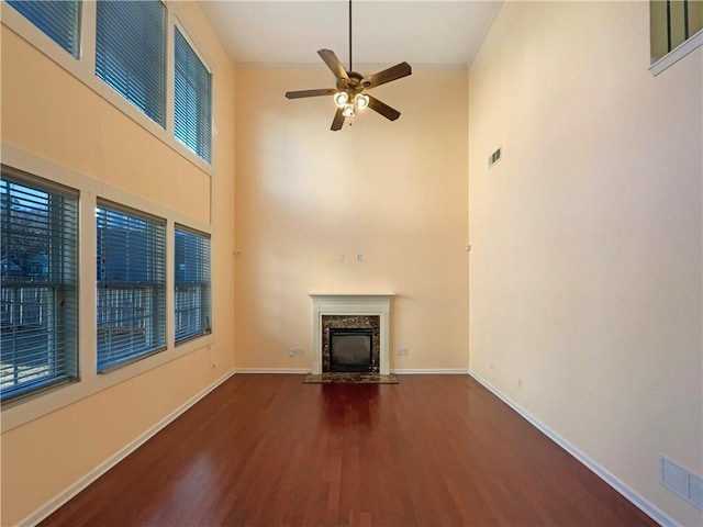 unfurnished living room with a towering ceiling, dark wood-type flooring, a premium fireplace, and ceiling fan