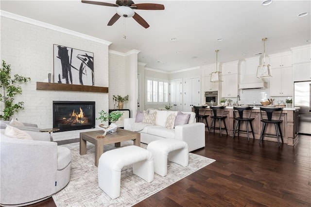 living room featuring a brick fireplace, ornamental molding, ceiling fan, dark wood-type flooring, and sink