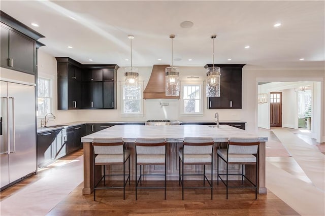 kitchen with pendant lighting, custom exhaust hood, stainless steel built in refrigerator, and a center island with sink