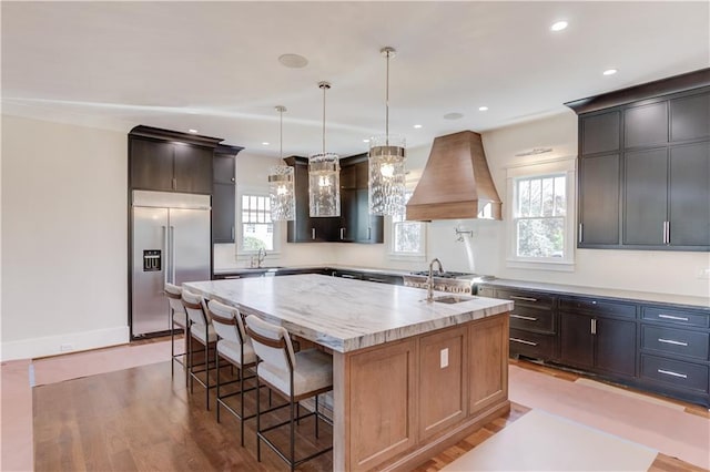 kitchen featuring stainless steel built in refrigerator, custom exhaust hood, hanging light fixtures, light stone countertops, and a center island with sink