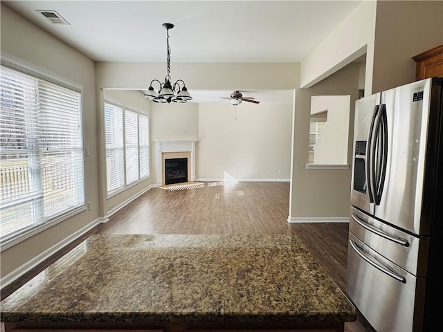 kitchen featuring stainless steel refrigerator with ice dispenser, ceiling fan with notable chandelier, dark wood-type flooring, pendant lighting, and dark stone countertops