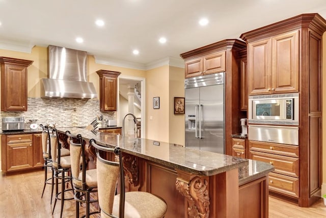 kitchen with wall chimney range hood, built in appliances, a warming drawer, crown molding, and light wood-style flooring