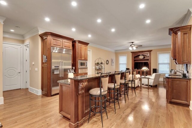 kitchen with built in appliances, a ceiling fan, dark stone counters, a breakfast bar area, and crown molding