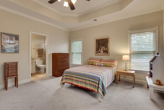 carpeted bedroom with a raised ceiling, ornamental molding, multiple windows, ensuite bath, and a ceiling fan