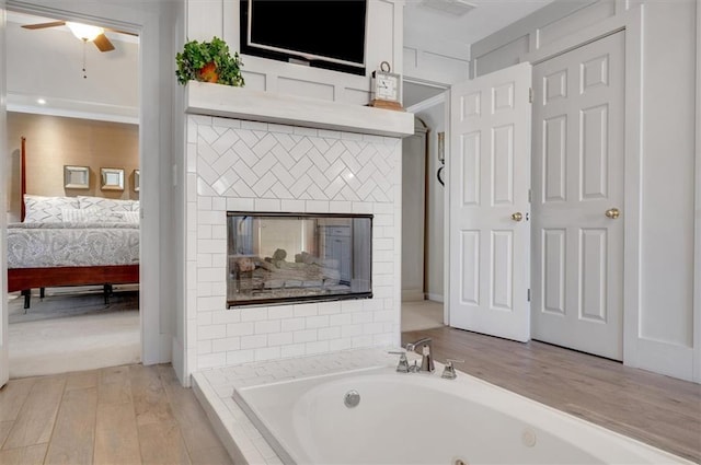 ensuite bathroom with ceiling fan, a multi sided fireplace, a whirlpool tub, ensuite bathroom, and wood finished floors