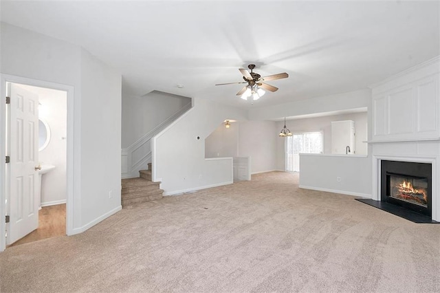 unfurnished living room featuring stairway, baseboards, light carpet, a glass covered fireplace, and ceiling fan with notable chandelier