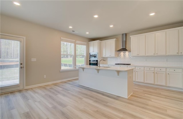 kitchen with wall chimney exhaust hood, a center island with sink, stainless steel microwave, oven, and white cabinets