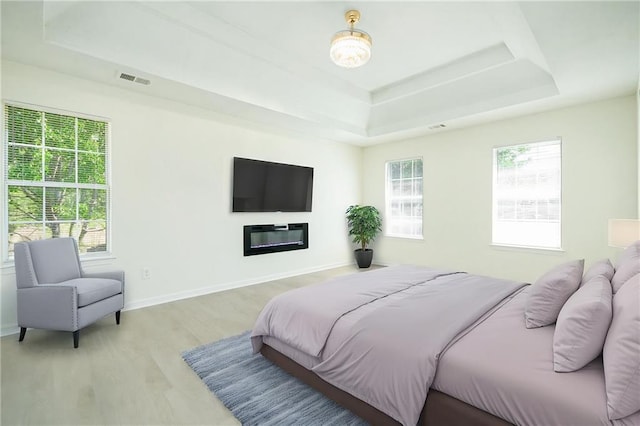 bedroom with light wood-style flooring, a raised ceiling, a glass covered fireplace, and baseboards