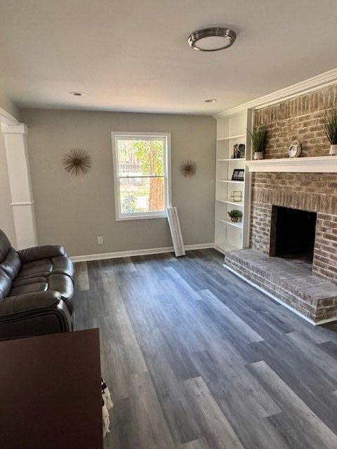 living room with a brick fireplace, built in shelves, baseboards, and dark wood-style flooring