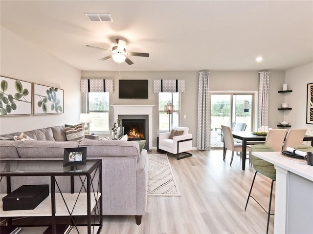 living room with light wood finished floors, visible vents, a ceiling fan, a warm lit fireplace, and baseboards