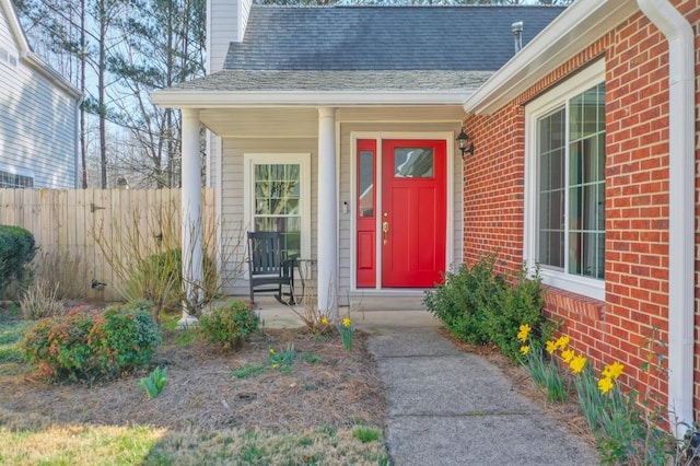 view of exterior entry with brick siding, a shingled roof, a chimney, and fence