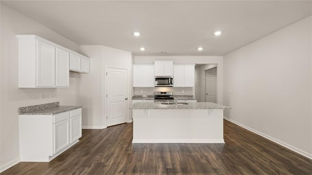 kitchen with light stone countertops, appliances with stainless steel finishes, white cabinets, dark wood-type flooring, and a center island with sink