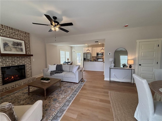 living room featuring light wood finished floors, ceiling fan, a brick fireplace, and baseboards