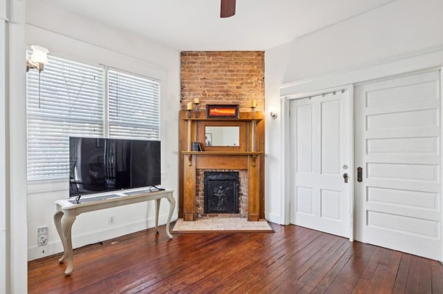 living room with ceiling fan, dark wood-style flooring, a brick fireplace, and baseboards