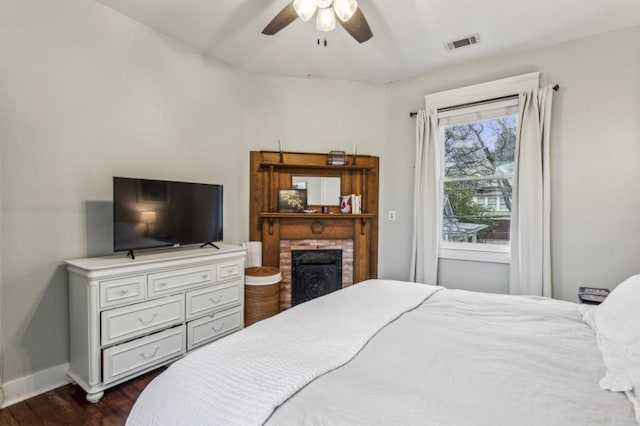 bedroom with a brick fireplace, dark wood-type flooring, and ceiling fan