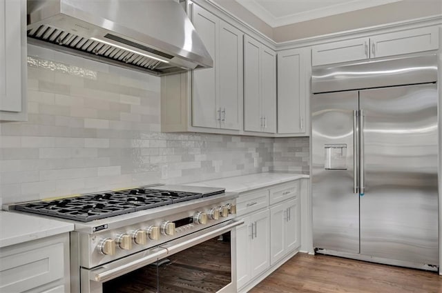 kitchen featuring high quality appliances, light wood-type flooring, decorative backsplash, light stone countertops, and wall chimney exhaust hood