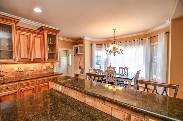 kitchen featuring brown cabinetry, ornamental molding, dark stone countertops, pendant lighting, and backsplash