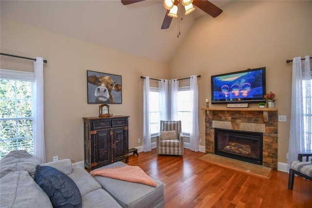 living area with baseboards, ceiling fan, wood finished floors, a stone fireplace, and high vaulted ceiling