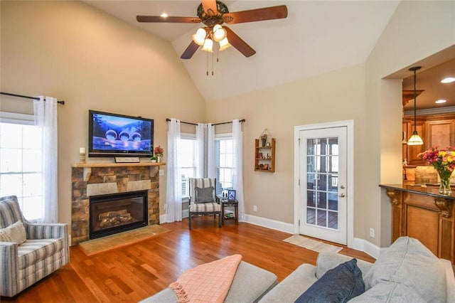 living area with ceiling fan, high vaulted ceiling, a stone fireplace, wood finished floors, and baseboards