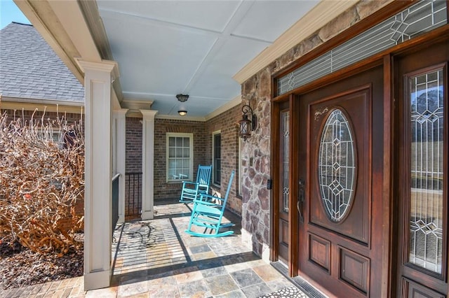 entrance to property featuring covered porch, brick siding, and roof with shingles