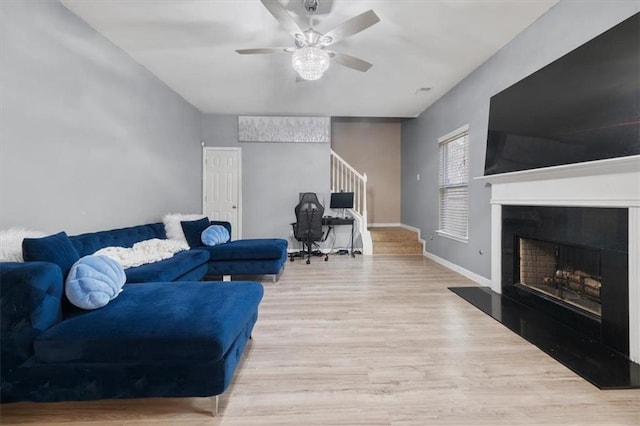 living room featuring a fireplace with raised hearth, a ceiling fan, wood finished floors, baseboards, and stairs